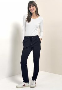 Černé zúžené kalhoty s nastavitelným pasem na šňůrku, v kombinaci s bílou košilí s dlouhým rukávem a bílými teniskami, stojící na neutrálním pozadí.