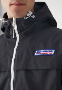 Mężczyzna ubrany w czarną kurtkę z białym zamkiem błyskawicznym i niebiesko-czerwoną naszywką z logo Napapijri na piersi, kaptur z białymi sznurkami.