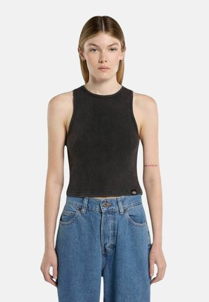 Donna con capelli biondi lisci che indossa un top corto nero senza maniche a coste e jeans blu a vita alta su uno sfondo semplice.