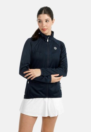 Vrouw in een marineblauwe jas met rits en een witte rok staat met gekruiste armen en kijkt iets opzij tegen een lichte effen achtergrond.