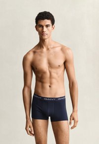 Junger Mann mit kurzen lockigen Haaren, der marineblaue GANT Boxershorts trägt, steht vor einem einfarbigen hellen Hintergrund.