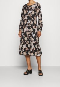 Schwarzes, langärmeliges Blumenkleid mit beigen und weißen Mustern, geraffter Taille und ausgestellt geschnittenem Saum; kombiniert mit schwarzen Sandalen mit Blockabsatz.