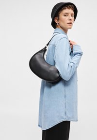 Μαύρη δερμάτινη τσάντα ώμου με καμπύλο σχήμα, με κλείσιμο φερμουάρ και αποσπώμενη λουρί, φορεμένη πάνω από ένα ανοιχτό τζιν πουκάμισο.