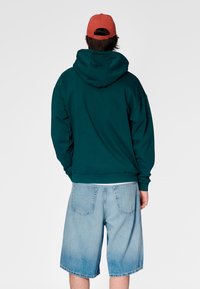 Türkiser Kapuzenpullover mit lockerem Schnitt und Rippbündchen, kombiniert mit hellblauen Denim-Shorts. Einfaches Design, keine sichtbaren Muster oder Akzente.