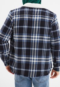 Camicia in flanella blu navy con motivo a quadri blu e bianco. Presenta un collo in sherpa crema, maniche lunghe e un orlo curvo.