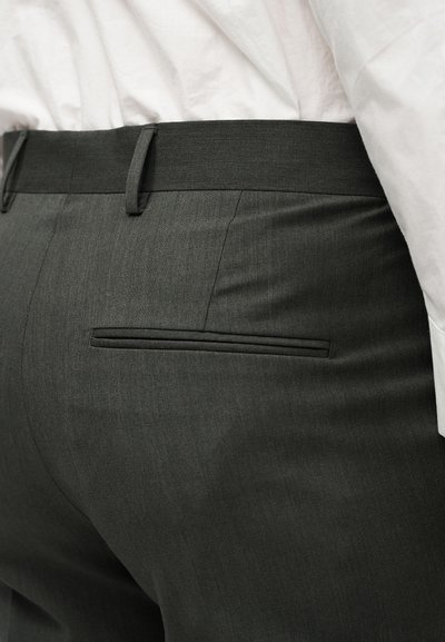 Κοντινό πλάνο ενός ατόμου που φοράει σκούρα γκρι παντελόνι με πίσω τσέπη με πατιλέτα και θηλιές για ζώνη, συνδυασμένο με λευκό πουκάμισο.