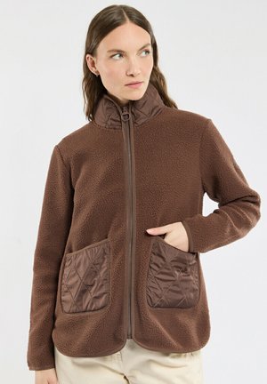 Femme portant une veste polaire marron avec un col montant et des poches matelassées, debout sur un fond uni clair, regardant sur le côté.