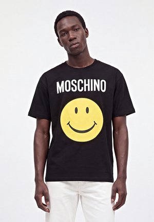 Jeune homme portant un t-shirt noir Moschino avec un grand motif de smiley jaune et un pantalon blanc sur un fond uni.