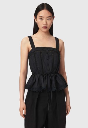 Femme aux cheveux noirs raides porte un haut péplum noir sans manches avec des boutons et des détails en dentelle, associé à un pantalon noir, fond neutre.