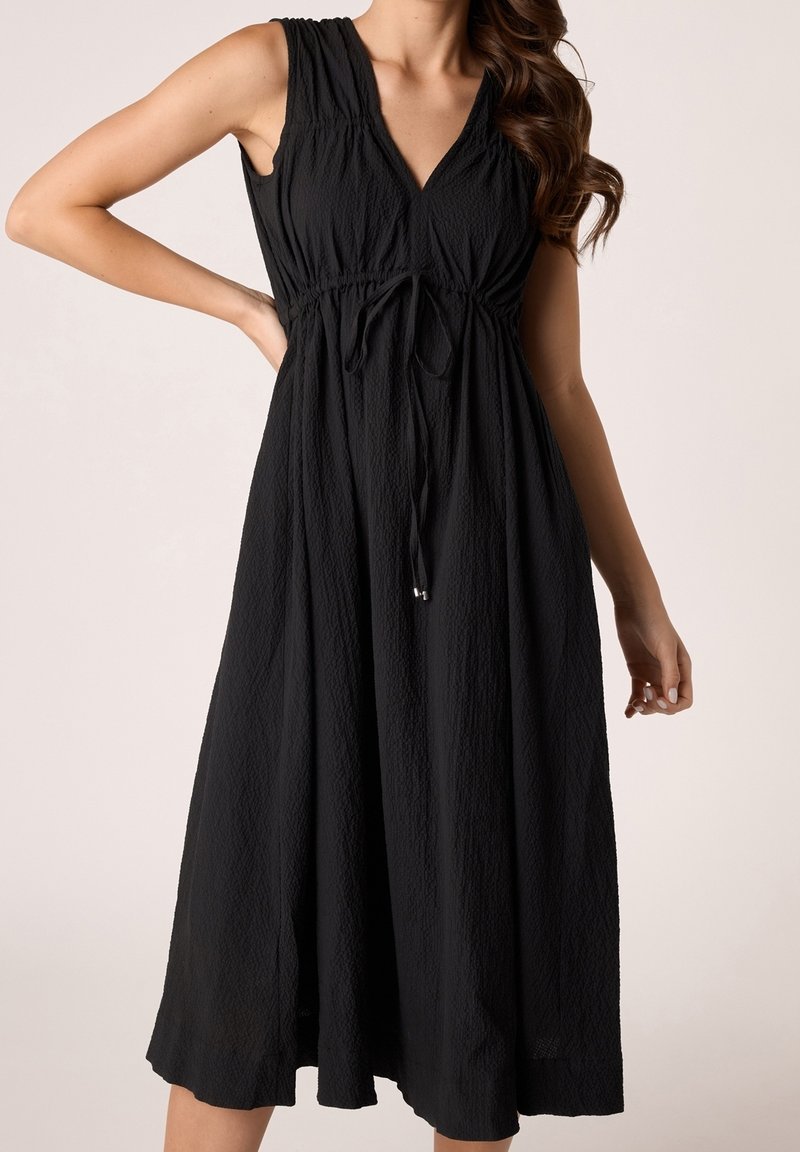 Robe midi noire texturée sans manches avec col en V et lien ajustable à la taille, portée par une femme aux cheveux ondulés.