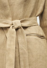 Μπεζ υφασμάτινη υφή δεμένη σε κόμπο γύρω από τη μέση ενός ενδύματος με μια κοντινή λεπτομέρεια σχισμής τσέπης.