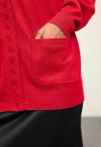 Cardigan en tricot rouge avec une rangée de boutons assortis et une poche latérale. Confectionné dans un matériau doux, il présente des bords côtelés aux poignets et à l'ourlet.