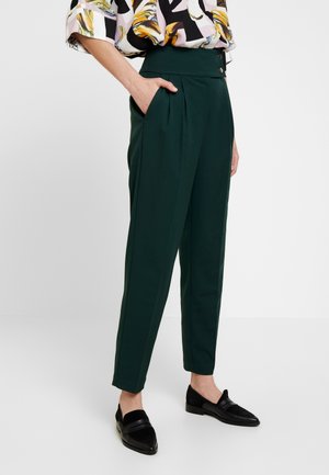 Henkilö, jolla on päällään tummanvihreät korkeavyötäröiset kapealahkeiset housut, joissa on laskokset, sivutaskut ja mustat loaferit, yhdistettynä kuvioituun paitapuseroon.