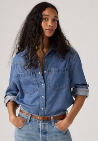 Lyseblå denimskjorte med knaplukning foran, to brystlommer og oprullede ærmer. Båret med et brunt læderbælte over blå jeans.