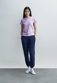 Ανοιχτό μωβ βαμβακερό T-shirt με στρογγυλή λαιμόκοψη, συνδυασμένο με παντελόνι φόρμας σε σκούρο μπλε. Λευκά αθλητικά παπούτσια ολοκληρώνουν την εμφάνιση. Ομαλή υφή ορατή.