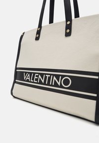 Canvas shoppingväska i beige med svarta läderdetaljer. Utrustad med "VALENTINO"-logotyp och dubbla svarta ränder. Messingnitar på handtagen för stöd.