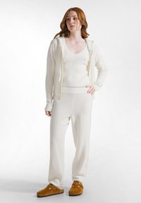 Conjunto de loungewear blanco de punto que incluye una camiseta sin mangas, un cárdigan con capucha y pantalones de corte holgado. Textura suave con detalles de canalé, acompañado de zapatillas en tono beige.