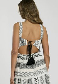 Vestido blanco con patrones geométricos negros, espalda abierta sujeta por lazos y acentos de flecos en la falda. Presenta tirantes anchos y un diseño fluido.