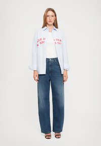 Chemise à rayures bleues et boutons, avec texte rouge, portée sur un t-shirt blanc et accompagnée de jeans larges bleus et de sandales noires.