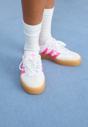 Sneakers brancos com riscas rosas e logótipo, biqueira de camurça, sola de borracha, combinados com meias brancas de cano curto com textura, sobre um fundo azul.