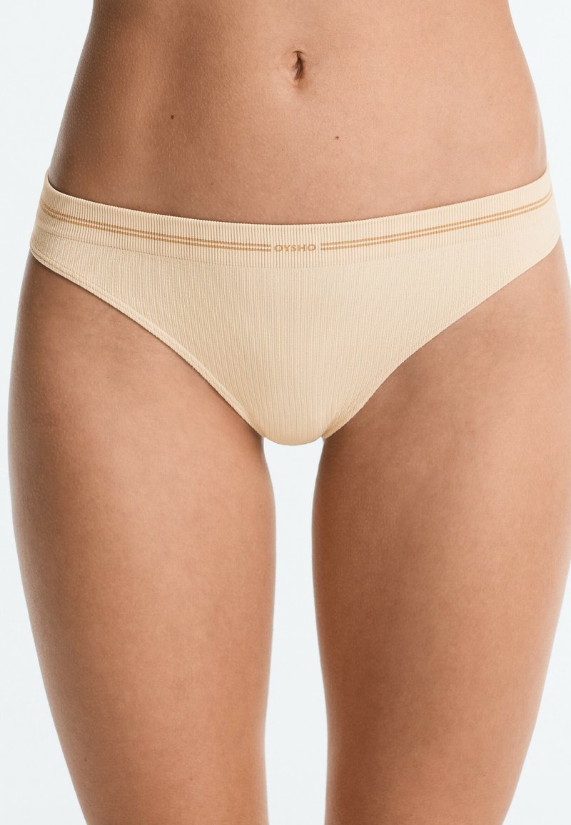 Personne portant des sous-vêtements côtelés beige clair, montrée de la taille au haut des cuisses, avec un logo et une discrète rayure près de la ceinture.