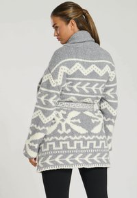 Grå och vit stickad cardigan med geometriska mönster, med en hög krage, vida ärmar och en lös passform, visad bakifrån.