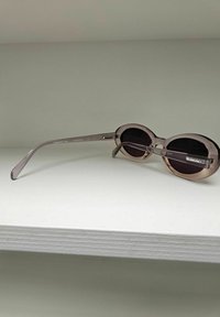 Lunettes de soleil rondes avec une monture translucide rose clair et des verres foncés. Fines branches s'amincissant vers les extrémités, portant la marque "KOMONO".