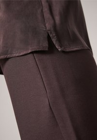 Haut brun à couches avec un dos légèrement plus long, fabriqué dans un matériau doux ; associé à un pantalon brun ajusté assorti, créant un look cohérent.