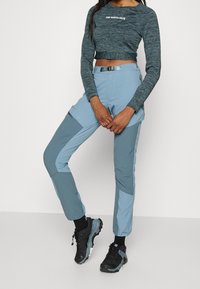 Ljusblå byxor med mörkblå accenter, tillverkade av ett mjukt, elastiskt material. En croppad grå topp med en vit logotyp. Svarta träningsskor.