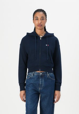 Hoodie court bleu marine avec fermeture éclair, présentant une poche avant et des cordons de serrage ajustables. Porté avec un jean en denim taille haute.