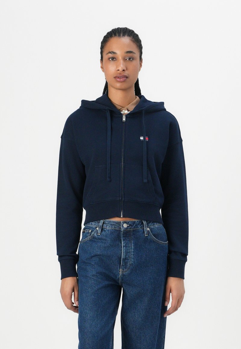 Hoodie corto di colore blu navy con zip, dotato di tasca frontale e lacci regolabili. Indossato con jeans in denim a vita alta.