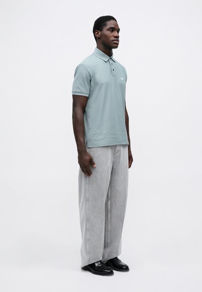 Ανοιχτό πράσινο βαμβακερό πόλο μπλουζάκι με τρία κουμπιά, συνδυασμένο με χαλαρούς γκρι τζιν παντελόνες και μαύρα παπούτσια. Το μοντέλο στέκεται στο προφίλ μπροστά σε λευκό φόντο.