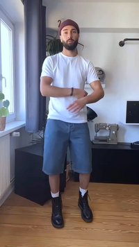 Moški, oblečen v klobuk, belo majico in jeans kratke hlače, stoji notri blizu okna, prilagaja svoj rokav, v ozadju pa je šivalni stroj.