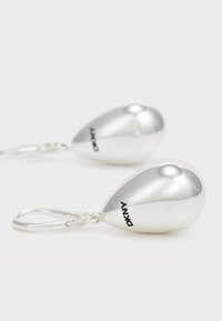 Silverfärgade tårformade örhängen med en glänsande yta och DKNY-logotyp, hängande från enkla krokar mot en neutral bakgrund.