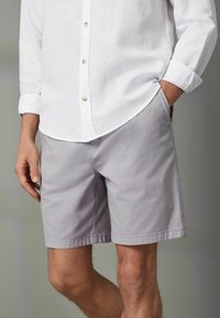 Hellgraue Shorts mit einer glatten Textur, ausgestattet mit Seitentaschen und einem schlichten Design. Getragen mit einem weißen Hemd mit Knöpfen.