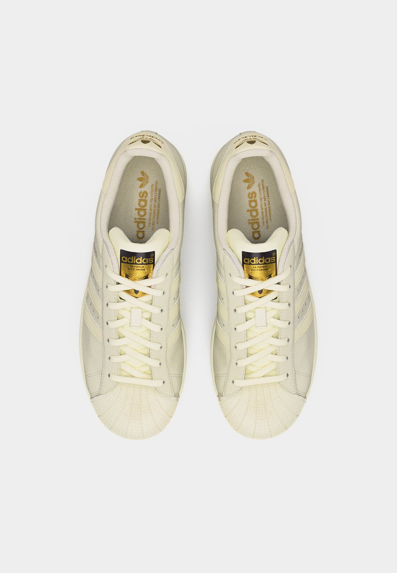 Baskets Adidas couleur crème avec embouts en forme de coquillage, logos Adidas dorés sur la languette et les semelles intérieures, et lacets blancs, vues de dessus.
