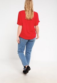 Blusa roja de mangas cortas con puños enrollados, combinada con jeans de color azul claro y calzado negro, vista desde atrás.