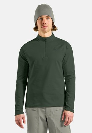 Dunkelgrüner Langarmpullover mit Reißverschlusskragen aus glattem Stoff. Kombiniert mit einer hellgrauen Strickmütze. Minimales Logodetail vorhanden.