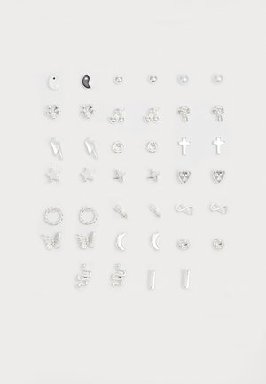 Conjunto de brincos de prata com várias formas: estrelas, corações, cruzes e símbolos como serpentes e borboletas, com uma mistura de texturas e acabamentos.
