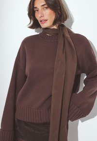 Femme aux cheveux bruns courts portant un pull en laine marron foncé et une écharpe assortie, se tenant contre un mur blanc avec la main sur la hanche.