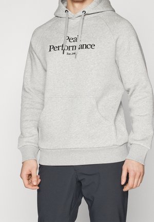 Jasnoszara bluza z kapturem z przednią kieszenią i ściągaczami, czarny napis "Peak Performance Est. 1986" na piersi, noszona z ciemnymi spodniami.