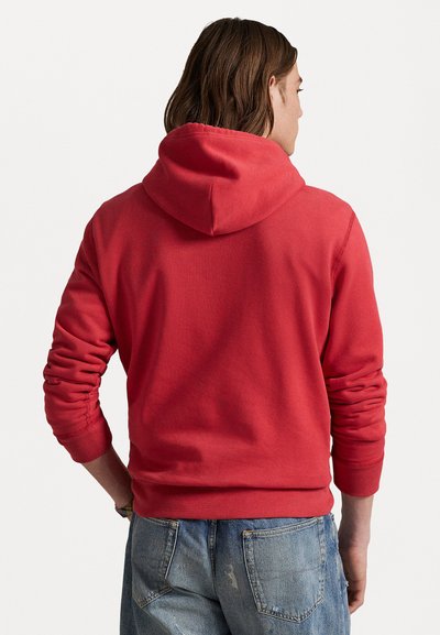 Sarkans hoodie ar mīkstu tekstūru, pieguļošiem manžetēm un josta. Aizmugurē ir vienkāršs dizains, un redzama kapuce. Zem tā ir uzvilkti džinsi.
