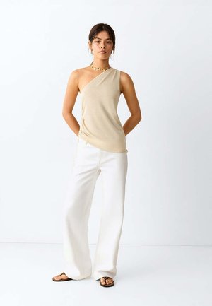 Mujer con top beige de un solo hombro, pantalones blancos de pierna ancha, sandalias negras y joyas doradas, posando frente a un fondo blanco liso.