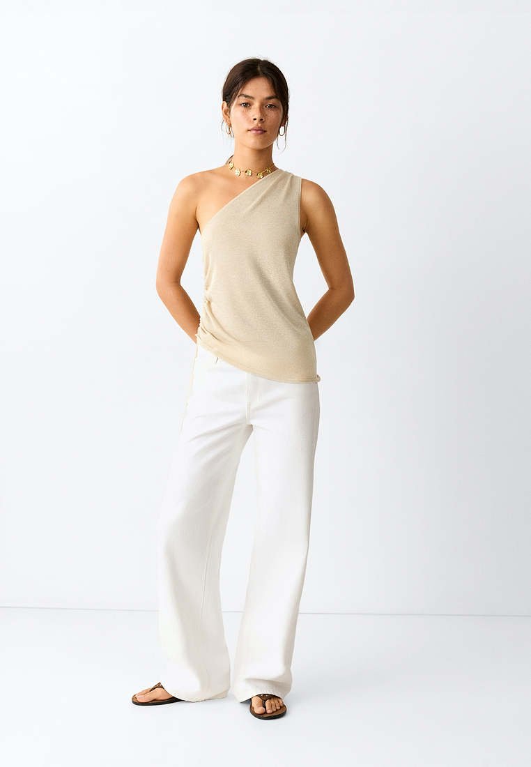 Femme portant un haut beige à une épaule, un pantalon blanc à jambes larges, des sandales noires et des bijoux en or, debout sur un fond blanc uni.