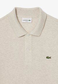 Beige Lacoste slim-fit pikétröja med en texturerad tyg, klassisk krage och en grön broderad krokodil-logotyp på vänster bröst.