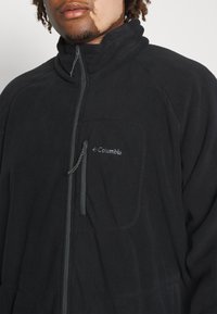 Giacca in pile nera con colletto alto, chiusura con zip e tasca sul petto. Caratterizzata da un branding discreto e un tessuto morbido e testurizzato.