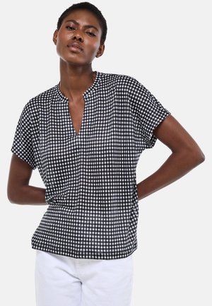 Femme portant un haut à manches courtes à carreaux noirs et blancs avec une encolure fendue et un pantalon blanc, debout les mains derrière le dos.