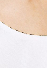 Vêtement blanc avec une texture lisse, doté d'un décolleté bordé de petites perles dorées pour l'accent. Aucun motif ou design supplémentaire.