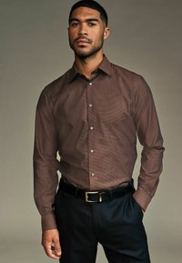 Cămașă maro, cu mâneci lungi și nasturi, cu un model subtil, asociată cu pantaloni negri și o curea lucioasă neagră cu o cataramă aurie.