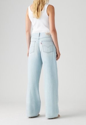 Kobieta widziana od tyłu, ubrana w jasnoniebieskie szerokie dżinsy i białą bezrękawnik na tle jasnego, jednolitego tła.